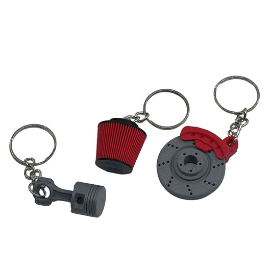 Three miniature car parts keychains on a white surface: a gray piston, a red and black air filter, and a gray brake rotor with a red brake caliper.